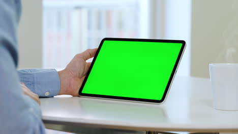 Man-in-blue-shirt-holding-tablet-with-green-screen,-white-table,-coffee-cup