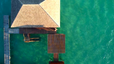 Mujer-Sentada-En-Un-Complejo-Privado-De-Villas-Sobre-El-Agua-Al-Amanecer-Mirando-Hacia-El-Agua-Azul-Turquesa,-Vista-Aérea-De-Arriba-Hacia-Abajo-Drone-Volando-Hacia-Arriba