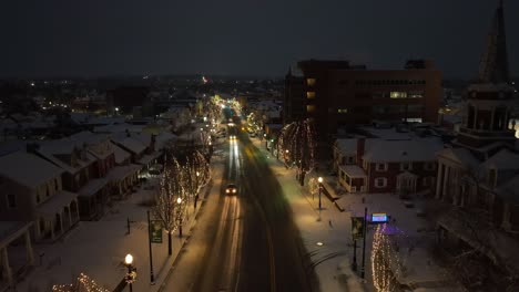 Nighttime-aerial-view-of-a-snow-covered-downtown-street-lined-with-holiday-lights,-glowing-warmly-under-a-dark-sky-in-the-USA