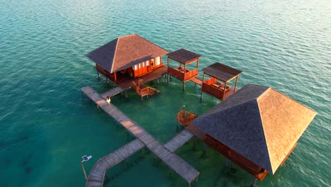 young-woman-walking-on-the-bridge-toward-an-overwater-villa-bungalow-at-sunrise,-aerial-view