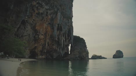 Olas-Tranquilas-Rompiendo-Suavemente-En-Una-Playa-Pintoresca-Junto-A-Un-Imponente-Acantilado-De-Piedra-Caliza-Con-Una-Cueva-En-Railay,-Tailandia