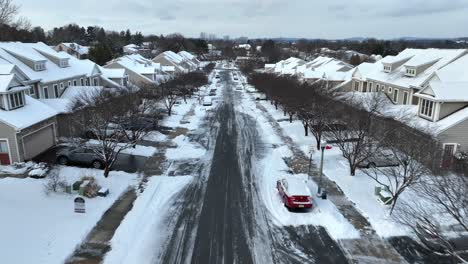 Aerial-view-of-a-snowy-suburban-street-lined-with-identical-houses,-cars-parked-on-driveways,-and-bare-trees