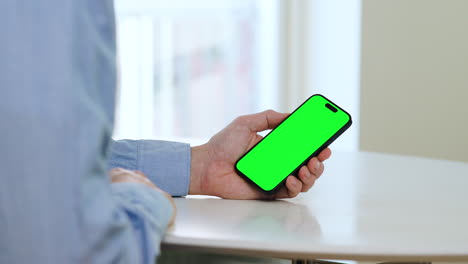 Green-screen-template-smartphone,-man-in-blue-shirt-sitting-by-white-table