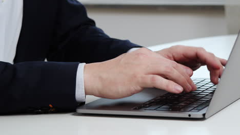 Hombre-Con-Chaqueta-Azul-Oscuro-Y-Camisa-Azul-Claro-Sentado-Junto-A-La-Mesa-Escribiendo-En-Una-Computadora-Portátil