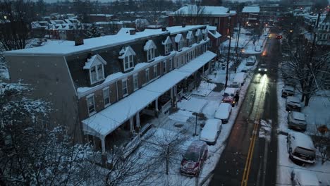 Early-evening-aerial-view-of-a-snowy-residential-street-lined-with-row-houses,-illuminated-by-street-lights-in-the-USA