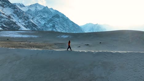 Aerial-View-Of-Male-Walking-Along-Sarfaranga-Cold-Desert-Ridge-With-Snow-Landscape-Mountains-In-Background