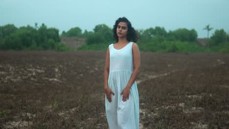 Woman-in-a-white-dress-standing-in-an-open-field-with-sparse-vegetation,-gazing-thoughtfully-into-the-distance