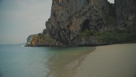 Waves-Gently-Breaking-On-Empty-Beach-Beside-Cliff-With-Cave-In-Railay,-Thailand