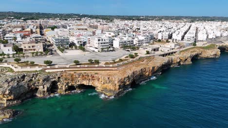 Vista-Aérea-Giratoria-Que-Revela-Lentamente-El-Escarpado-Acantilado-Frente-Al-Mar-Con-Polignano-A-Yegua-Al-Fondo,-Italia