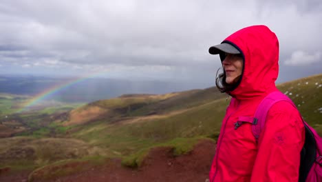 Surprised-woman-hiker-pointing-to-a-rainbow-in-a-mountainous-landscape,-Wales