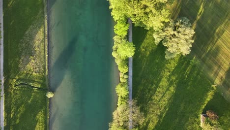 Bird-eye-view-from-above-of-clean-road-surrounded-by-green-grass-and-plant-trees-on-sunny-day,-river