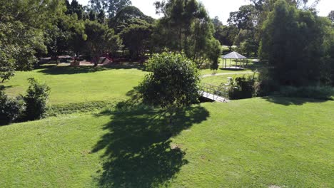 Drone-flying-low-towards-a-small-tree-in-a-green-park-also-showing-a-children's-playground-in-Australia