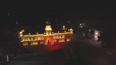 Toma-Aérea-De-Un-Dron-De-Un-Edificio-Histórico-De-La-Torre-Del-Reloj-Iluminado-Durante-La-Noche-En-La-Ciudad-De-Gwalior-En-Madhya-Pradesh,-India