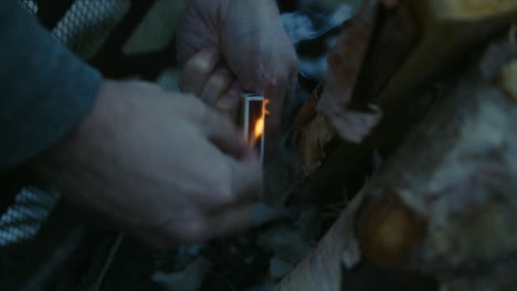 Man-striking-match-on-matchbox-to-light-fire-at-base-of-teepee-logs-in-pit