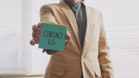 Un-Joven-Vestido-Con-Trajes-Formales-Mostrando-Notas-Adhesivas-Con-Texto-Contáctanos-A-La-Cámara