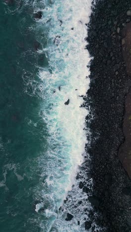 „Drohnenaufnahmen-Von-Einem-Schwarzen-Steinstrand-Von-Oben,-Der-In-Den-Himmel-Aufsteigt,-Während-Wellen-über-Die-Steine-Darunter-Fließen