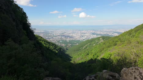 Beautiful-Dronie-shot-of-City,-as-seen-from-a-lush-green-mountain-forest,-on-a-sunny-day