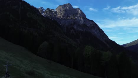 Person-Mit-Schwarzer-Jacke-Sitzt-Im-Schatten-Neben-Dem-Gipfelkreuz-Mit-Berglandschaft-Im-Hintergrund-Mit-Schneebedecktem-Gipfel-An-Einem-Sonnigen-Tag-In-Österreich,-Europa