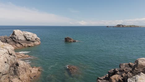 Drone-flight-from-rocky-shoreline-out-into-bay-on-bright-calm-day