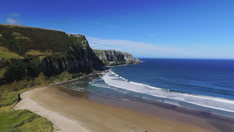 Aerial-view-of-Purakaunui-Bay-Scenic-Reserve-along-the-Catlins-Coast,-showcasing-the-stunning-coastline-with-dramatic-cliffs,-sandy-beach,-and-the-vast-expanse-of-blue-ocean-under-a-clear-sky