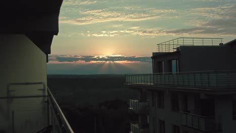 Atemberaubender-Blick-Auf-Den-Sonnenuntergang-Von-Einem-Modernen-Balkon-Mit-Heiterem-Abendhimmel,-Sonnenstrahlen,-Die-Durch-Die-Wolken-Brechen,-Und-Friedlicher-Stadtlandschaft