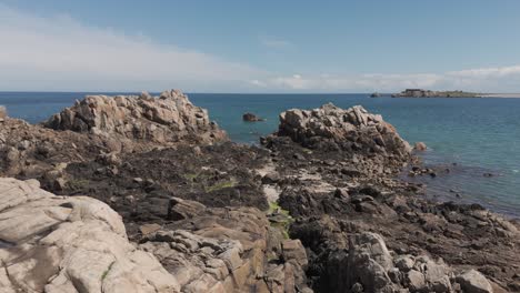 Drone-flight-at-low-tide-over-rocky-foreshore-with-rockpools-and-birds-to-inlet-and-views-across-bay-on-bright-sunny-day-in-Guernsey