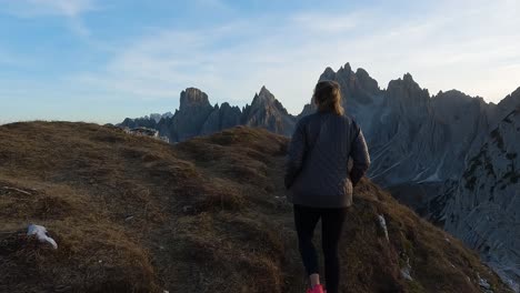 Rear-tracking-follows-woman-hiking-as-sun-sets-below-Dolomites,-backlit-mountain-vista