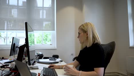 Blonde-Haired-Woman-Sitting-At-Desk-In-Office-Working-Looking-At-Monitor-And-Drinking-From-Mug