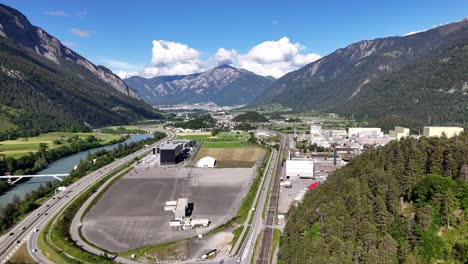 Domat-ems,-switzerland-with-industrial-area,-river,-and-mountains-on-a-sunny-day,-aerial-view