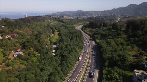 Aerial-view-of-the-road-passing-by-the-nature-side-black-sea-with-cars-and-trucks-passing-by-among-green-meadows-sunny-weather-sun-weather-afternoon-time