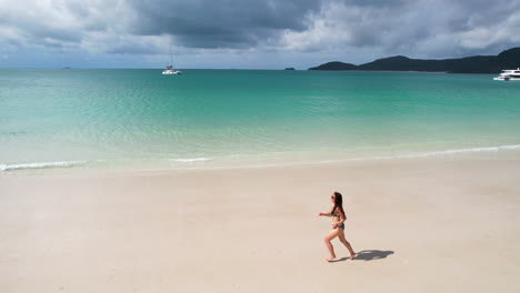 Aerial-View-of-Young-Happy-Woman-in-Bikini-Running-on-White-Sand-Beach-by-Turquoise-Sea-on-Exotic-Island