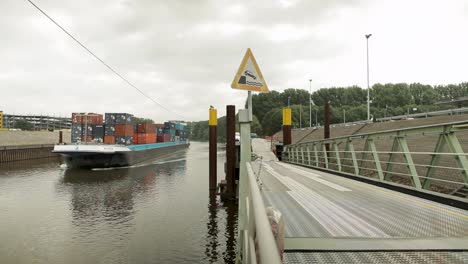 Cargo-ship-loaded-with-containers-floats-by-on-a-river,-viewed-from-a-pedestrian-bridge-with-a-warning-sign