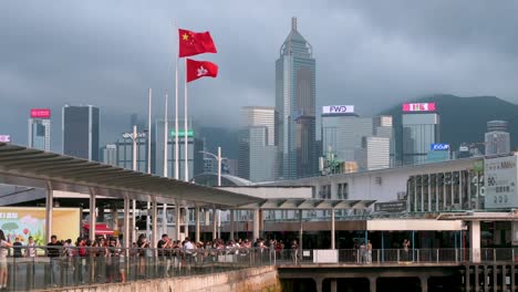 A-Lo-Largo-Del-Paseo-Marítimo,-Las-Banderas-De-Hong-Kong-Y-China-Ondean-Con-Gracia,-Con-Rascacielos-Alzándose-Detrás.