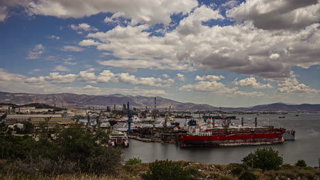 Toma-De-Tiempo-De-Nubes-Moviéndose-Sobre-Colinas-En-El-Fondo-Con-El-Puerto-De-Elefsina-Y-El-Sitio-Industrial-Petrolero-Helénico-En-Primer-Plano-En-Atenas,-Grecia