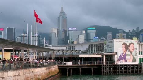 Fluttering-in-the-breeze,-flags-of-Hong-Kong-and-China-stand-tall-at-Victoria-Harbour-and-pier,-with-skyscrapers-in-view