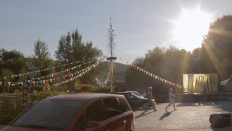 Sunlit-outdoor-market-scene-with-colorful-banners,-people-walking,-cars-parked