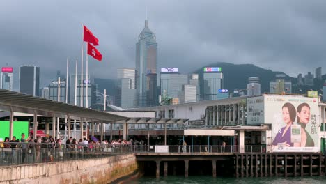 Banderas-De-Hong-Kong-Y-China-Ondeando-Al-Viento-En-El-Puerto-Y-El-Muelle-De-Victoria,-Con-Rascacielos-Visibles-Al-Fondo