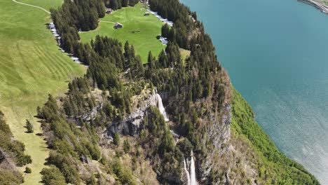 Aerial-view-of-Seerenbach-Falls-cascading-down-a-cliff-into-lush-green-surroundings-with-nearby-fields-and-the-blue-waters-of-Walensee-in-Amden,-Betlis,-Switzerland