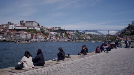 Menschen-Sitzen-Am-Flussufer-Von-Gaia-Und-Blicken-Auf-Den-Fluss-Und-Nach-Porto-Mit-Der-Dom-Luís-I-Brücke-Im-Hintergrund