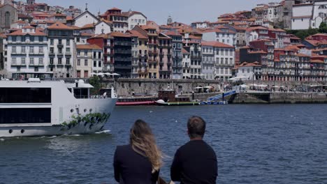 Couple-enjoying-the-riverside-view-at-Porto,-with-cruise-passing-on-the-background-on-a-sunny-day