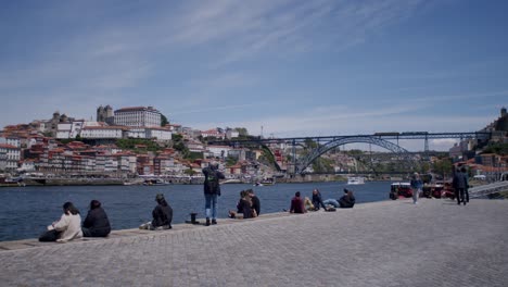 People-sitting-and-walking-on-the-riverside-of-Gaia-with-Dom-Luis-Bridge-on-the-background-on-a-sunny-morning