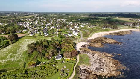 Coastal-village-with-white-houses,-green-fields,-and-rocky-shoreline-on-a-sunny-day,-aerial-view