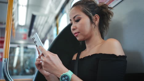 Woman-with-tied-back-hair,-wearing-a-black-off-shoulder-top,-is-engrossed-in-her-smartphone-while-riding-a-train-focusing-on-her-concentrated-expression-and-the-modern-interior-of-the-train