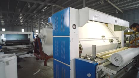 Worker-operates-modern-textile-machinery-in-a-Pakistani-factory,-processing-white-fabric-rolls