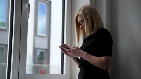 Frau-Im-Schwarzen-Kleid,-Die-Tagsüber-Am-Fenster-Einer-Modernen-Wohnung-Ihr-Smartphone-Benutzt