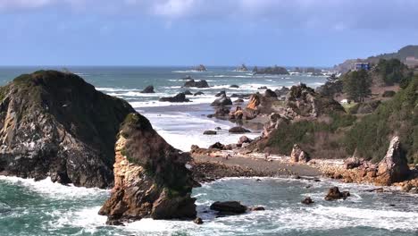 Discover-the-beauty-of-a-rocky-beach-with-a-distant-house-in-this-left-dolly-aerial-shot,-showcasing-dramatic-coastal-rocks,-rolling-waves,-and-serene,-secluded-charm