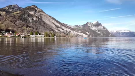 Un-Encantador-Perro-Blanco-Recupera-Un-Palo-De-Madera-De-Las-Aguas-Del-Lago-Walensee-En-Suiza,-Con-El-Majestuoso-Telón-De-Fondo-De-Los-Alpes.