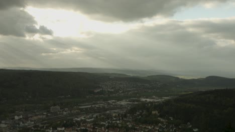 A-lush-valley-with-rays-of-sunlight-piercing-through-stormy-clouds,-highlighting-a-small-town,-aerial-view