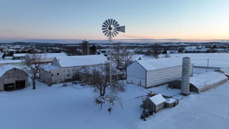 Die-Morgendämmerung-Erhellt-Einen-Schneebedeckten-Bauernhof-Mit-Scheunen,-Einem-Silo-Und-Einer-Klassischen-Windmühle-Und-Verkörpert-Eine-Ruhige-Ländliche-Winterszene