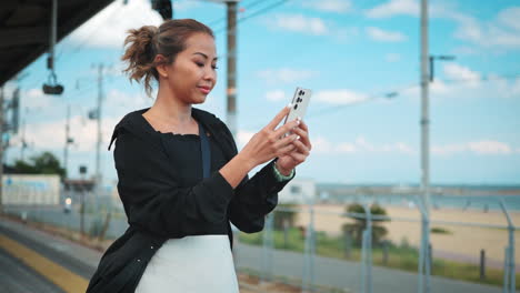 Woman-dressed-in-a-black-top-and-white-skirt,-focuses-on-her-smartphone-while-waiting-at-an-outdoor-train-platform